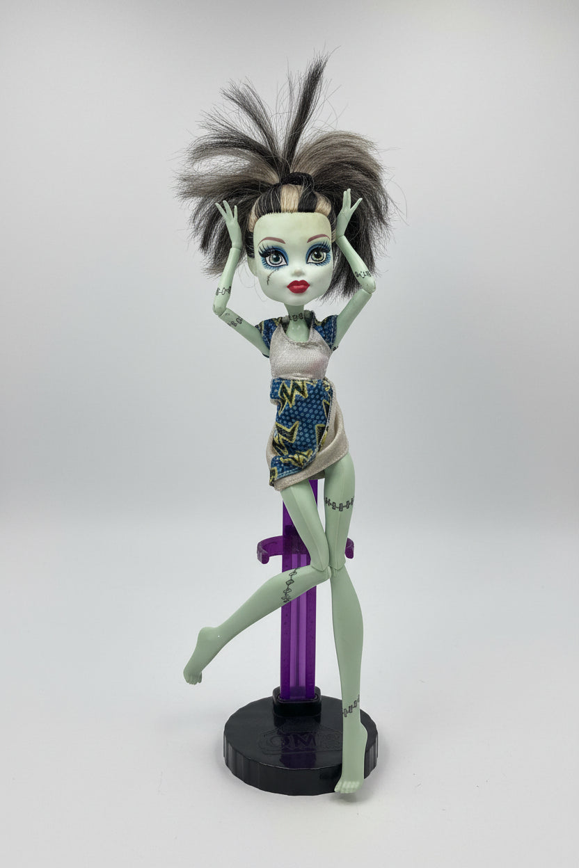 Monster High doll with spiky hair and colorful dress on a pink surface with blinds in the background
