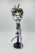 Monster High doll with spiky hair and colorful dress on a pink surface with blinds in the background